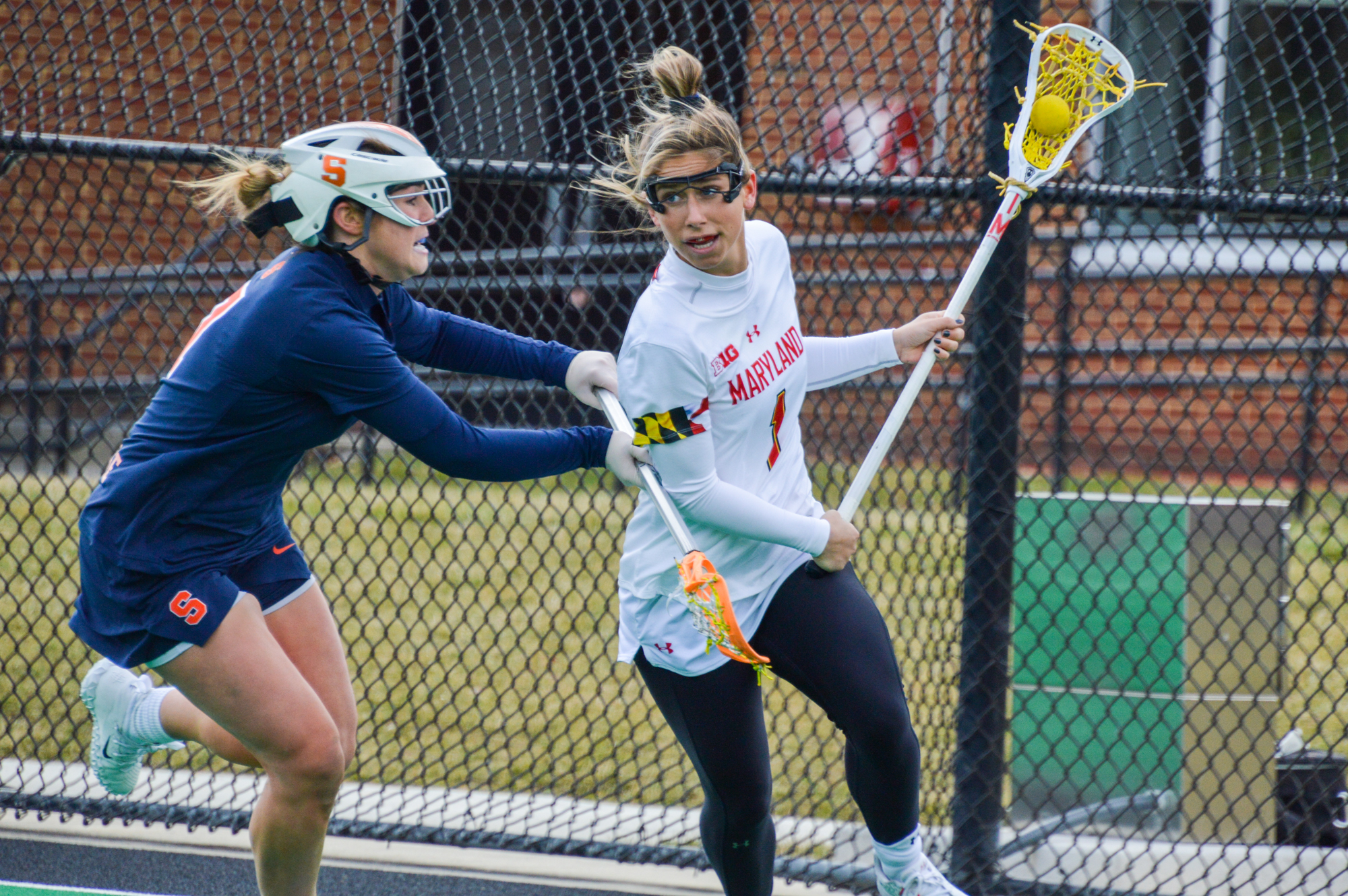 Woman in a white jersey with a lacrosse stick and ball running past a woman in a blue jersey with a lacrosse stick